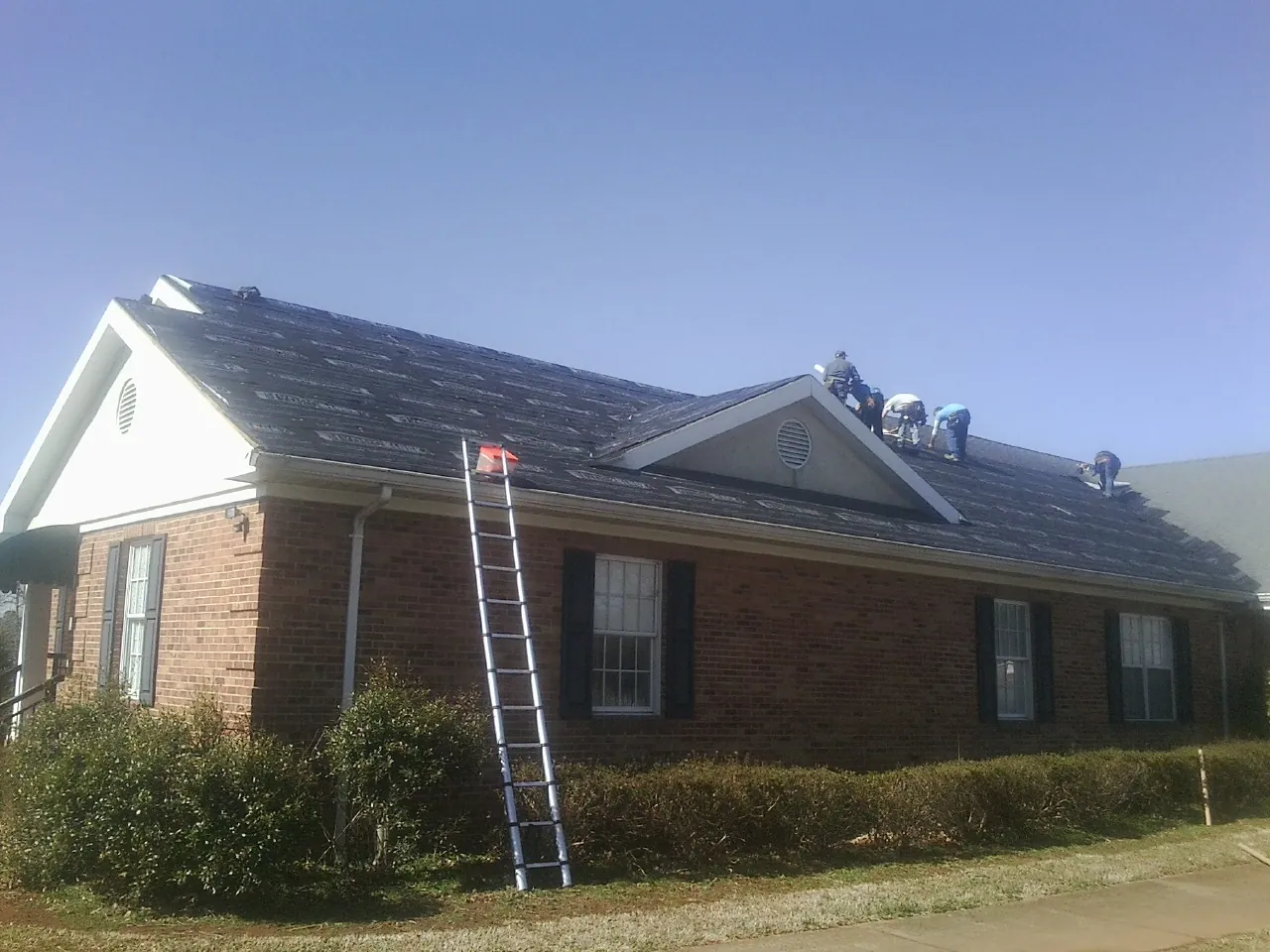 Crew tearing shingles off a brick ranch roof with a ladder set — Emergency Roof Repair work by our crew in Clio